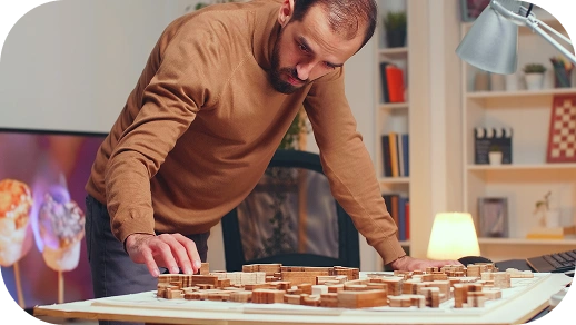 Een man in een bruine trui bekijkt aandachtig een gedetailleerd architectonisch model op een tafel in een gezellige, goed verlichte kamer met boekenplanken en een bureaulamp.